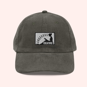 May include: An olive green corduroy baseball cap with a curved peak. The cap has a rectangular patch with the words "DANGEROUS CREATURE" and a silhouette of a person reading.