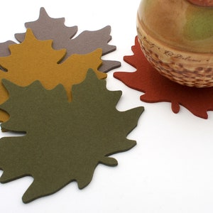 May include: Set of four felt leaf-shaped coasters in brown, yellow, green, and red. The coasters are arranged in a stack with a brown ceramic pitcher on top of the red coaster.