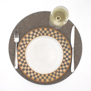 Puede incluir: Un mantel individual de fieltro marr&oacute;n con un plato blanco con un borde a cuadros negro y marr&oacute;n. Un tenedor y un cuchillo de plata est&aacute;n colocados en el mantel individual, y una copa de vino blanco est&aacute; sobre el mantel individual.