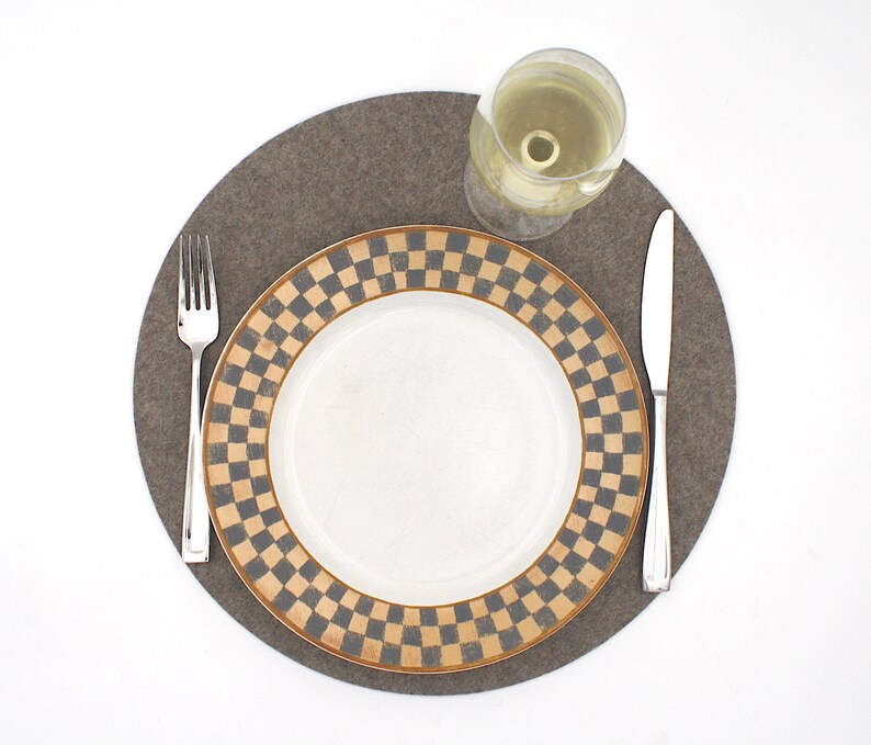 Puede incluir: Un mantel individual de fieltro marr&oacute;n con un plato blanco con un borde a cuadros negro y marr&oacute;n. Un tenedor y un cuchillo de plata est&aacute;n colocados en el mantel individual, y una copa de vino blanco est&aacute; sobre el mantel individual.