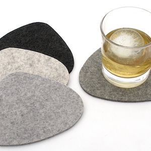 May include: A set of four felt coasters in shades of gray and black. One coaster is under a glass of whiskey with a large ice sphere. The coasters are pebble-shaped and made of a soft, textured material.