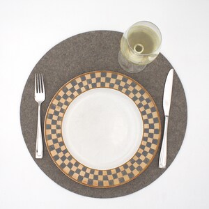 Puede incluir: Un mantel individual de fieltro gris con un plato blanco con un patr&oacute;n de cuadros negros y dorados. Un vaso de vino blanco y dos cubiertos de plata est&aacute;n en el mantel individual.