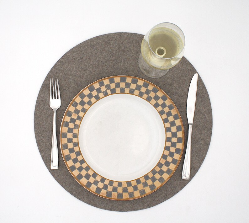 Puede incluir: Un mantel individual de fieltro gris con un plato blanco con un patr&oacute;n de cuadros negros y dorados. Un vaso de vino blanco y dos cubiertos de plata est&aacute;n en el mantel individual.