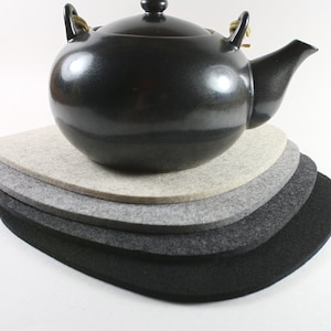 May include: Set of four felt coasters in various shades of gray, with a black ceramic teapot resting on top. The coasters are oval shaped and have a slightly raised edge.