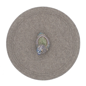 May include: A round gray felt coaster with concentric circles. A ceramic duck with blue, green, and white paint is on top of the coaster.