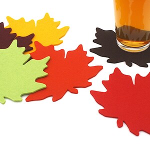 May include: Five felt maple leaf coasters in different colors: brown, green, red, orange, and black. A glass of amber liquid sits on the black coaster.