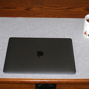 May include: A gray felt desk mat with a laptop, phone, mouse, and mug on a wooden desk. The laptop is closed, displaying the Apple logo. A white mug with a black and white design sits next to a white mouse.