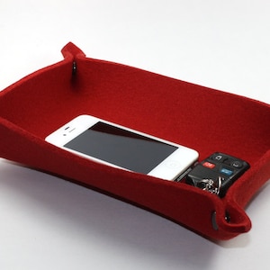 May include: A red felt tray with a rectangular shape and folded corners secured by dark gray snaps. Inside the tray are a white smartphone and a black car key fob. The tray is designed for organizing small items.