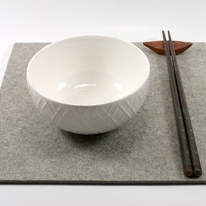 May include: A white ceramic bowl with a textured design sits on a gray felt placemat with a pair of wooden chopsticks and a wooden leaf-shaped chopstick rest.