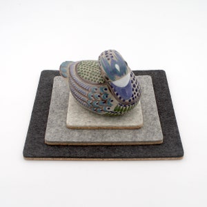 May include: A ceramic duck figurine with blue, green, and white details rests on three square felt coasters in shades of gray and black. The duck is perched on the smallest, light gray coaster.