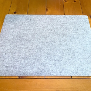 May include: A rectangular desk mat made of light gray felt material. The mat features a flat, uniform surface with a subtle mottled texture throughout, characterized by a blend of light and dark gray fibers, and has rounded corners.