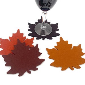 May include: A set of four felt drink coasters shaped like maple leaves in shades of dark brown, burnt orange, and mustard yellow. One dark brown coaster supports a clear glass stemware piece with a dark liquid and a visible etched crest.