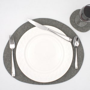May include: A gray felt placemat with a white plate, fork, knife, and spoon. The placemat is oval shaped and has a smooth texture.