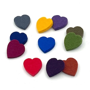 May include: A collection of eleven small, flat, heart-shaped felt cutouts in various colors including grey, yellow, purple, dark blue, bright blue, magenta, red, olive green, brown, and orange, arranged in a scattered pattern on a white surface.