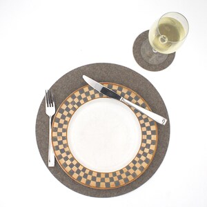 Puede incluir: Un mantel individual de fieltro marr&oacute;n con un patr&oacute;n a cuadros en beige y marr&oacute;n. Un plato blanco con un borde a cuadros beige y marr&oacute;n se coloca encima del mantel individual. Un tenedor y un cuchillo de plata se colocan en el plato. Una copa de vino blanco se coloca sobre un posavasos de fieltro marr&oacute;n.