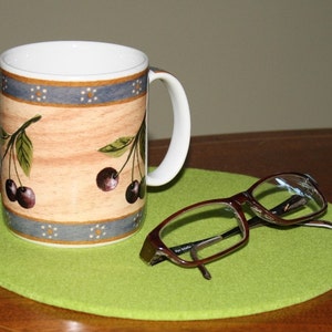 May include: A white ceramic mug with a brown, blue, and white cherry design. The mug is sitting on a green coaster. A pair of brown eyeglasses is lying on the coaster.