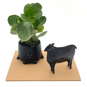 May include: A matte black ceramic planter with four small feet and a simple white smiley face graphic, containing a green plant with rounded leaves, positioned next to a solid black metal sheep figurine on a flat tan rectangular base.