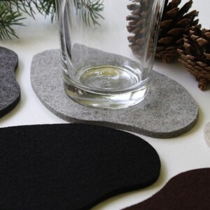 May include: Five felt coasters in different colors, including gray, black, beige, and brown. The coasters are shaped like irregular ovals and are arranged on a white surface. A glass is sitting on one of the gray coasters.