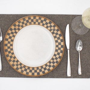 May include: A brown felt placemat with a white plate with a black and gold checkered border. A silver fork, knife, and spoon are placed on the placemat. A clear glass with a green liquid is on the placemat.