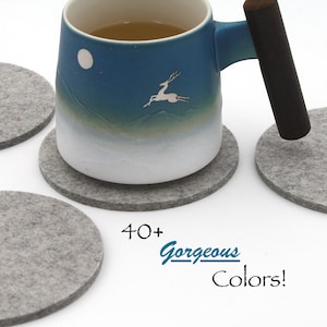 May include: A ceramic mug with a blue-to-white gradient, a white moon, and a white deer silhouette, featuring a dark brown wooden handle. The mug sits on a round grey felt coaster, surrounded by three additional grey felt coasters. Text reads 40+ Gorgeous Colours!
