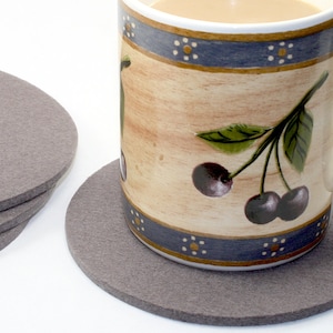 May include: A set of four gray felt coasters with a ceramic mug with a cherry design sitting on top of one of the coasters.