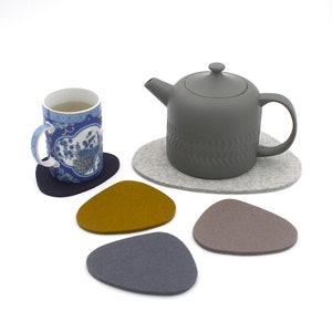 May include: Set of five felt coasters in gray, brown, blue, and yellow. The coasters are shaped like pebbles and are designed to protect surfaces from heat and scratches.