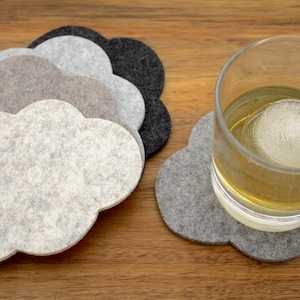 May include: Set of five felt coasters in gray, light gray, and black. The coasters are shaped like clouds and are arranged in a stack. A glass of whiskey with an ice cube is sitting on one of the coasters.