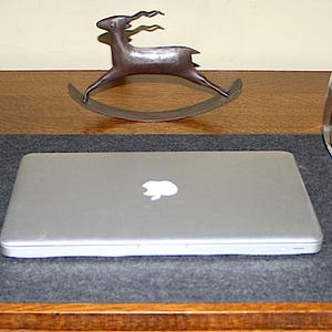 May include: A gray laptop computer with a silver case and a white Apple logo is on a gray desk mat. A silver phone is on the desk mat to the left of the laptop. A glass of water is on the desk to the right of the laptop. A metal deer sculpture is on the desk behind the laptop.