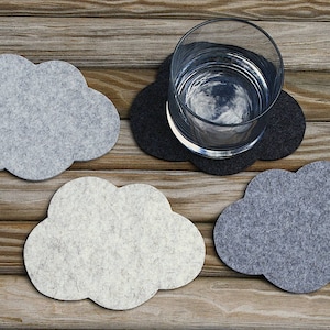 May include: Four gray and white felt cloud-shaped coasters on a wooden surface. One coaster has a clear glass with water on top.