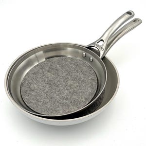May include: Two stainless steel frying pans, one nested inside the other. The top pan has a long, curved handle. A gray felt protector sits inside the top pan. The pans are on a white background.