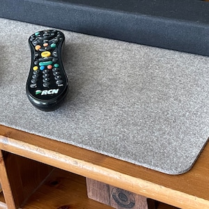 May include: A gray felt TV remote control organizer mat with two black remote controls on top. The mat is on a wooden surface.