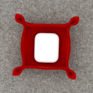 May include: A vibrant red felt tray, square in shape with rounded corners, containing a white wireless earbud charging case. The tray features four decorative, curved accents at each corner.