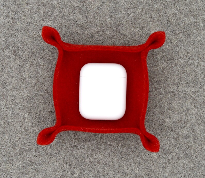 May include: A vibrant red felt tray, square in shape with rounded corners, containing a white wireless earbud charging case. The tray features four decorative, curved accents at each corner.