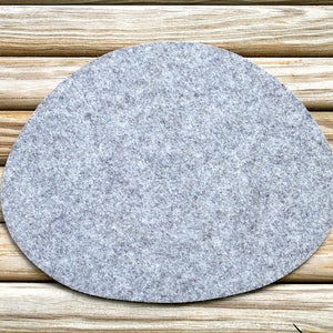 May include: A light gray, oval-shaped felt coaster. The coaster is set on a background of light brown wooden logs. The felt has a textured appearance.