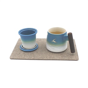 May include: A ceramic tea set featuring a blue and white gradient mug with a wooden handle and a matching tea infuser. The mug has a deer illustration. The set rests on a gray felt mat, perfect for tea lovers.