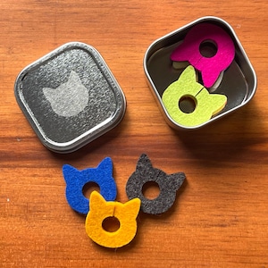 May include: A collection of colorful felt cat-shaped items. The image shows a silver tin with a cat silhouette, and several cat shapes in blue, yellow, and gray. Two cat shapes are inside a tin, in pink and green.