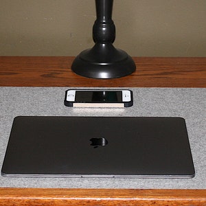 May include: A grey felt desk mat with a laptop, phone, and mouse. A red leather tray, a coffee mug with an apple design, and a set of keys are also on the desk. A black desk lamp is centered above the laptop.