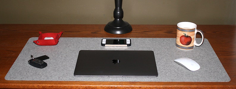 May include: A grey felt desk mat with a laptop, phone, and mouse. A red leather tray, a coffee mug with an apple design, and a set of keys are also on the desk. A black desk lamp is centered above the laptop.