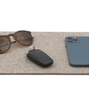 May include: A light gray felt tray with rounded corners holds a pair of brown sunglasses, a black car key fob, and a blue smartphone. The items are arranged on the tray against a white background.