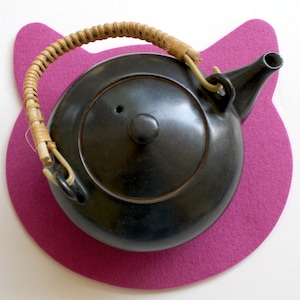May include: A black ceramic teapot with a woven bamboo handle sits on a pink felt cat-shaped coaster.