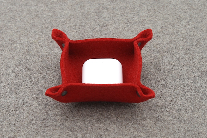 May include: A red felt tray with folded corners and small metal rivets. Inside the tray is a white rectangular object. The tray is on a grey felt surface. The tray is designed to hold small items.
