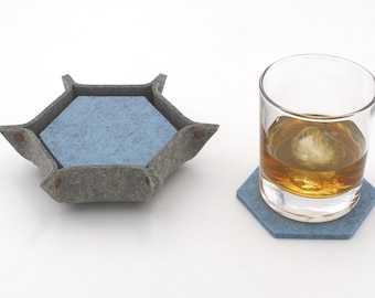 Puede incluir: Una bandeja hexagonal de fieltro gris con un interior de fieltro azul y un vaso de líquido ámbar con un cubito de hielo sobre un posavasos hexagonal de fieltro azul.