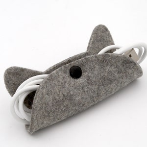 May include: A grey felt cord keeper shaped like a cat's head with a black button closure. A white charging cable is coiled inside the keeper.