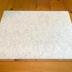 May include: A rectangular light gray felt mat with a mottled texture and rounded corners. The surface of the mat features a dense, fibrous appearance with subtle variations in tone throughout, creating a consistent and uniform look across the piece.