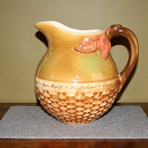 May include: A ceramic pitcher with a brown and tan glaze, a woven basket pattern, and a leaf-shaped handle. The pitcher has the words "Rome Beauty", "Red Delicious", and "Empire" written on it.