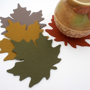 May include: Four felt maple leaf coasters in shades of brown, gold, and green. A brown ceramic pitcher sits on top of a red coaster.