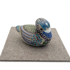 May include: A ceramic duck figurine with blue, green, and white painted details. The duck is sitting on a gray felt surface.