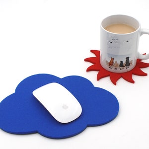 May include: A white computer mouse rests on a blue cloud-shaped mousepad. A white mug with a dog illustration sits on a red sun-shaped coaster. The mug contains a light brown liquid.