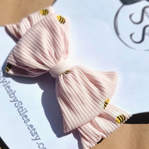 May include: A pink ribbed headband with a large bow and a bee print. The headband is on a white background with the text "sunlitstylesbystyles.etsy.com".