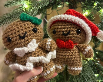 May include: Two crocheted gingerbread man ornaments. One wears a green bow and white trim, the other a Santa hat and red bow tie. Both have black eyes and white accents. The ornaments are brown and white.